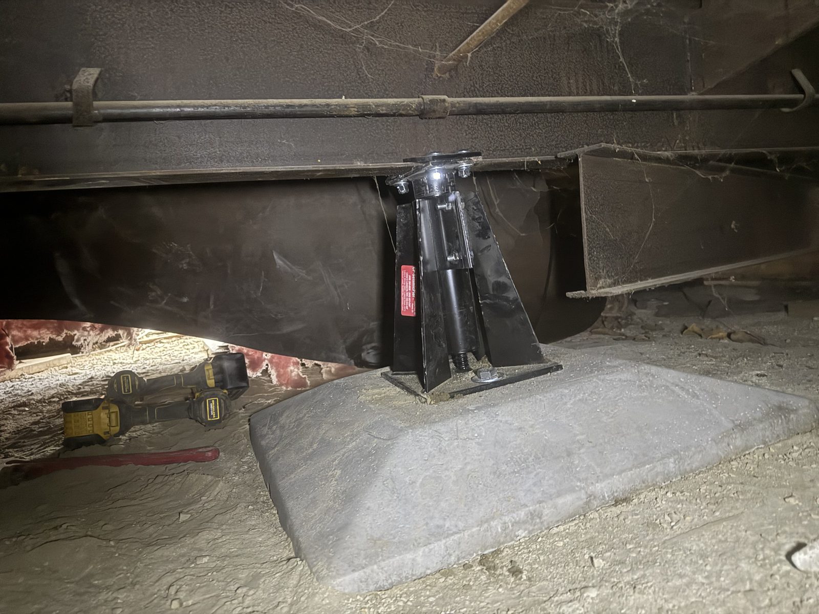 Wider view of an engineered pier on a concrete footing pad carrying the steel I-beam frame of a manufactured home, mid-installation.