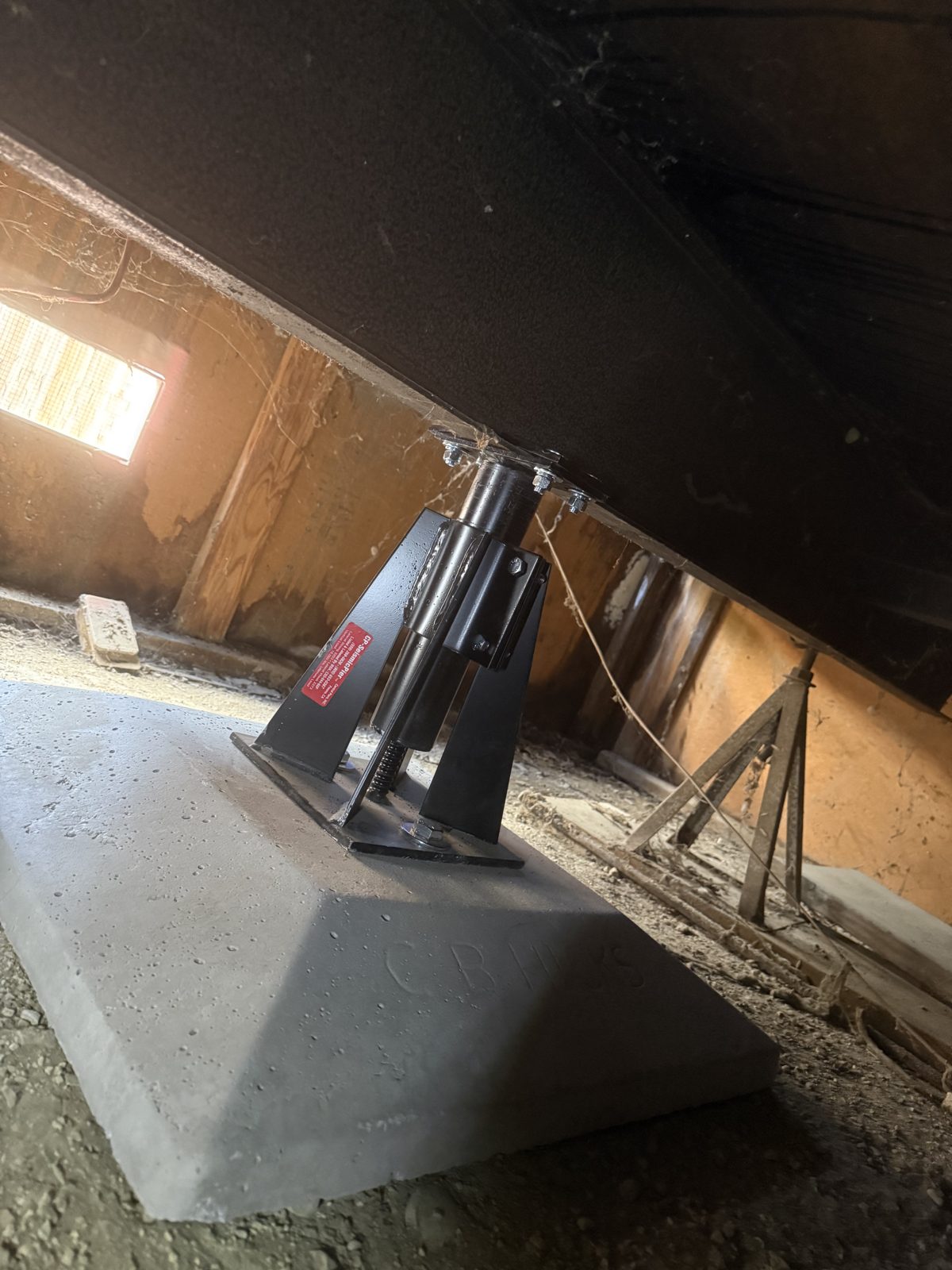 An engineered adjustable steel seismic pier on a concrete footing, bolted to the steel chassis beam of a manufactured home.